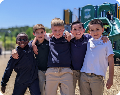 Four boys smiling and posing together on a playground, surrounded by swings and slides under a clear blue sky.