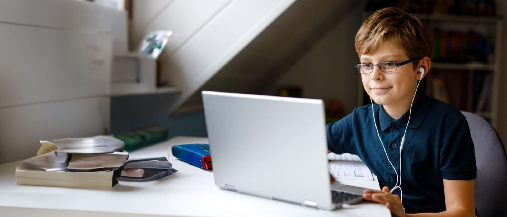 A young boy wearing glasses and headphones sitting at a desk with a laptop in front of him.